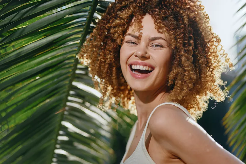 smiling woman with curly hair enjoys sunny tropical outdoors, happy young adult in casual white top surrounded by green palm leaves and natural light