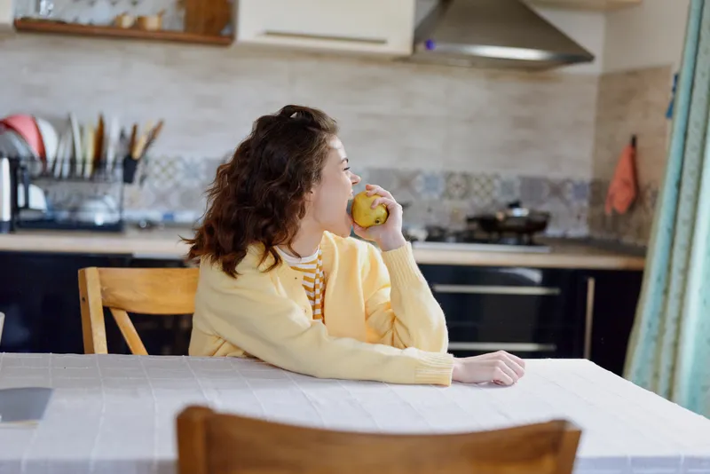 Cheerful woman indoors at home, sitting at a table in a bright kitchen, enjoying an apple, soft natural light, relaxed pose, casual style, warm colors, candid moment in calm, sunny mood