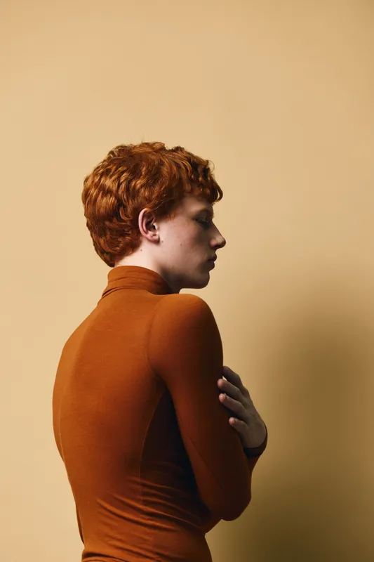 Redhead woman in brown turtleneck sweater standing sideways against beige background with folded arms and thoughtful expression on face in minimalistic studio setting.