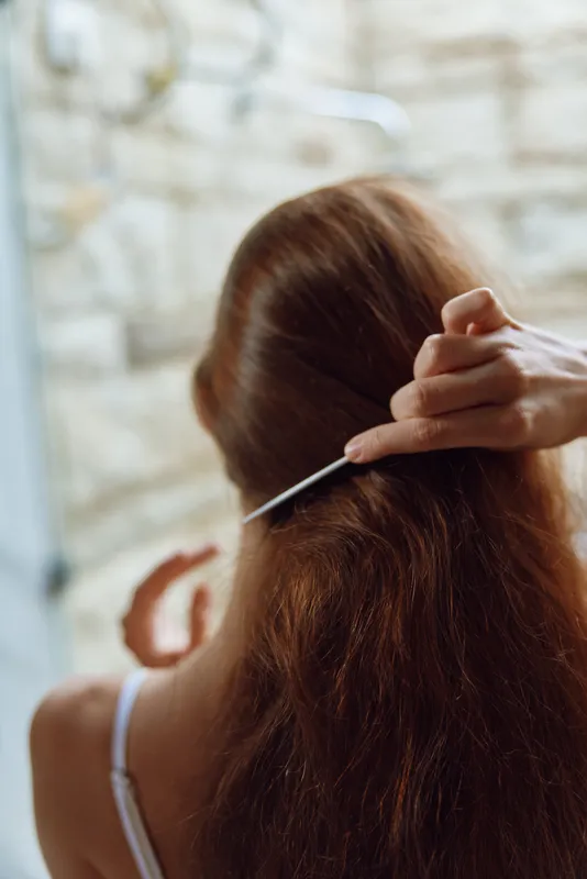 Woman combing long chestnut hair with white comb indoors near textured wall, casual morning routine, natural light highlighting soft strands, intimate self-care moment.