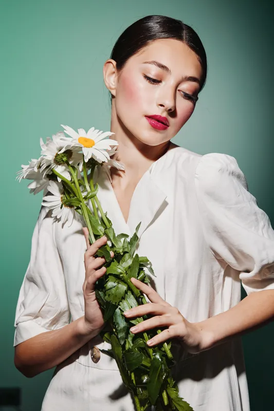 Young woman in white holding daisies with closed eyes. Soft moody lighting highlights her radiant skin and mauve lips. Beauty, creative fashion, floral elegance.