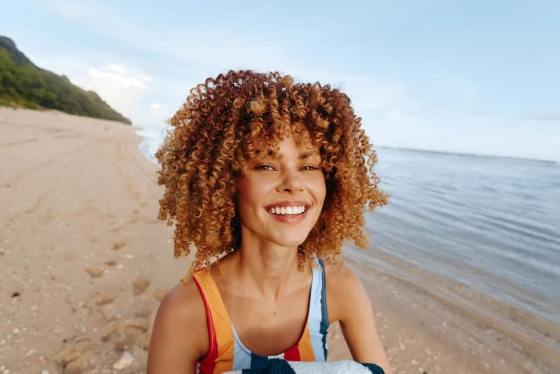 Smiling woman with curly hair standing on a sandy beach near the ocean. Bright daylight and clear sky create a cheerful and relaxed summer atmosphere.