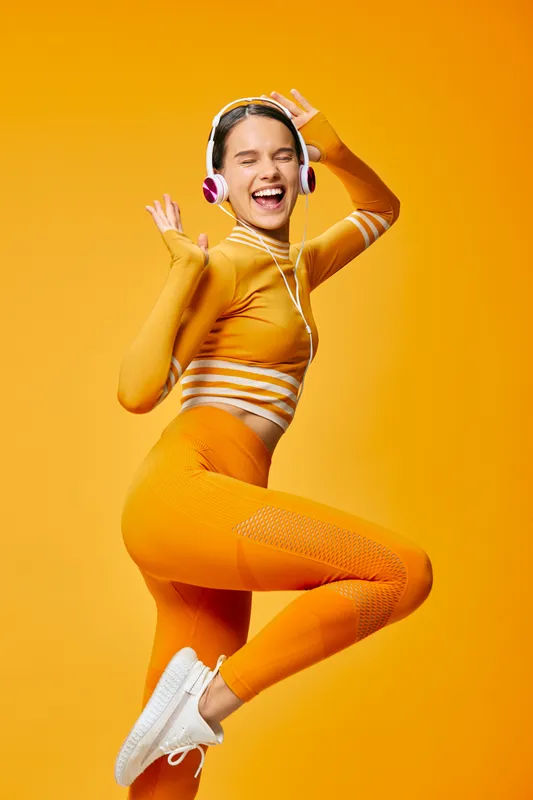 Fitness woman in bright orange athletic wear leaps with headphones against a bold orange backdrop, showing high energy, movement, and dance-inspired workout in a vibrant lifestyle scene.