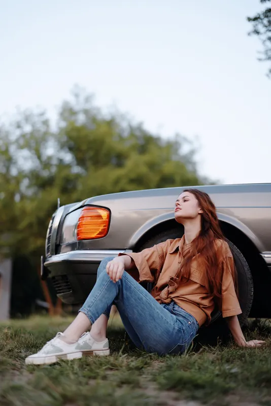 Relaxed young woman with long red hair in casual outfit sitting beside vintage car in natural setting, conveying a sense of freedom and serenity.