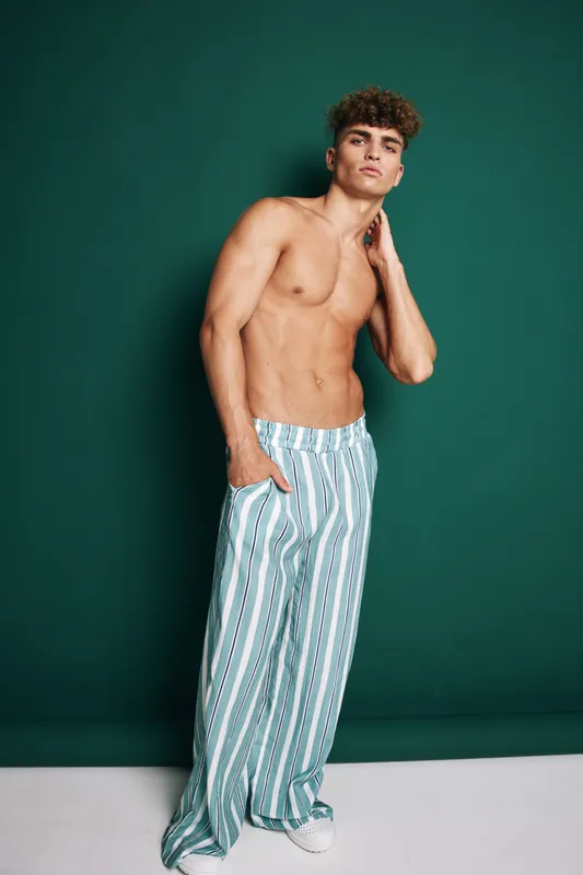 Young man with curly hair posing shirtless in striped wide pants and white sneakers against green background, showing confident and relaxed posture.