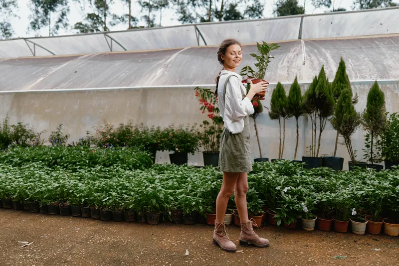 Young woman in a greenhouse holding plants, wearing a white blouse and green skirt, enjoying nature with greenery in the background and warm earthy tones