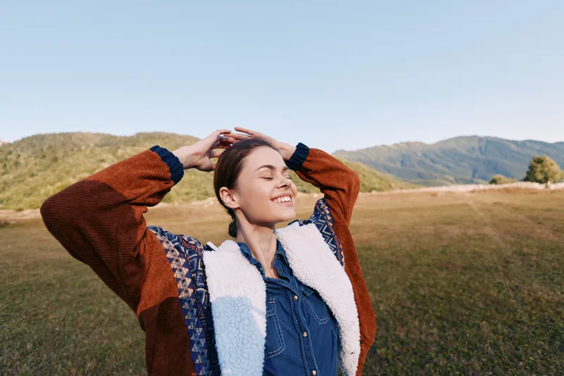 Woman smiling in nature with mountains and meadow background outdoors, feeling happiness and relaxation; portrait of a young woman in cozy jacket enjoying sunlight and fresh air.