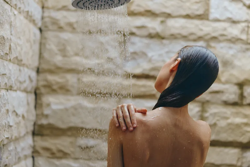Woman showering outdoors with water pouring from showerhead, wet hair, natural stone background, peaceful and refreshing morning routine atmosphere.