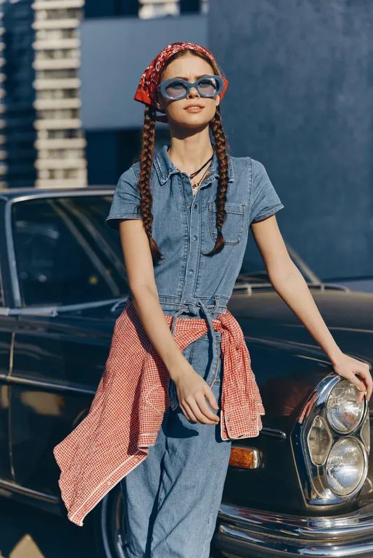 stylish woman with braids wearing sunglasses and retro outfit stands near classic black car in sunny urban setting with blue wall background