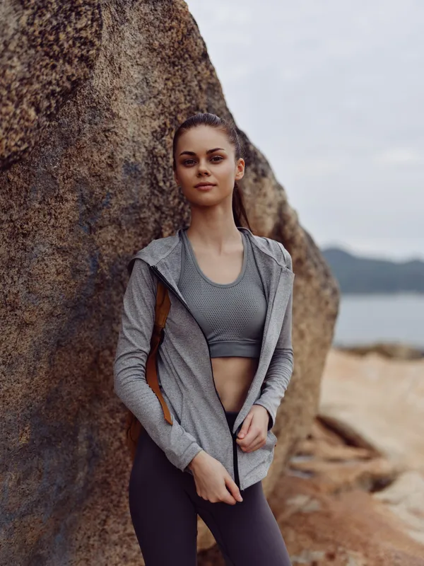 Young female model in activewear expressing confidence stands by a rocky beach background, promoting fitness and healthy lifestyle concepts