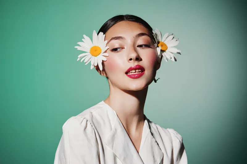 Young woman with fresh designed floral hairstyle featuring white daisies wears glossy lipstick and a white shirt against bright green. Beauty creative fashion editorial style shot.