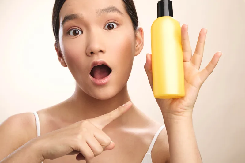Asian woman with wide eyes points at a yellow bottle, conveying surprise and curiosity in a bright studio setting, inviting interest in a personal care product and usage.