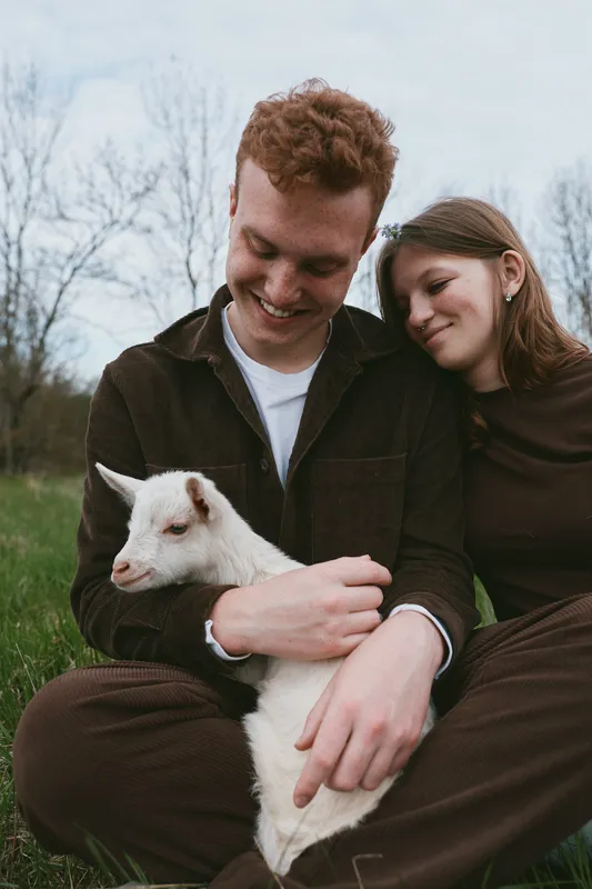 A couple shares a gentle moment with a cheerful goat kid in a grassy meadow, embracing outdoors, showing rural lifestyle and affectionate bond, serene skies, casual clothing, warm interaction,