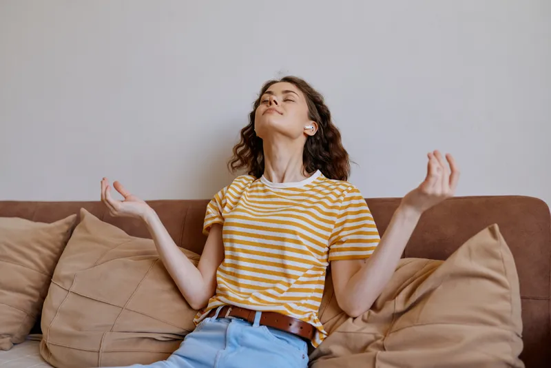 A woman relaxes on a sofa at home, meditating with closed eyes and gentle posture. Bright casual setting, soft cushions, warm light. Concept for selfcare, mindfulness, relaxation, and comfortable