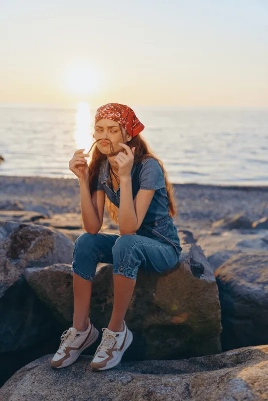 young woman sitting on rocks by the sea wearing a red headscarf and denim overalls at sunset with a peaceful and contemplative mood