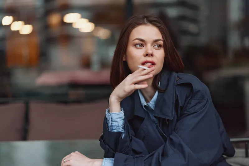 Thoughtful young woman sitting indoors at a table, looking away, wearing a dark jacket and light denim shirt, soft lighting, casual atmosphere, pensive mood