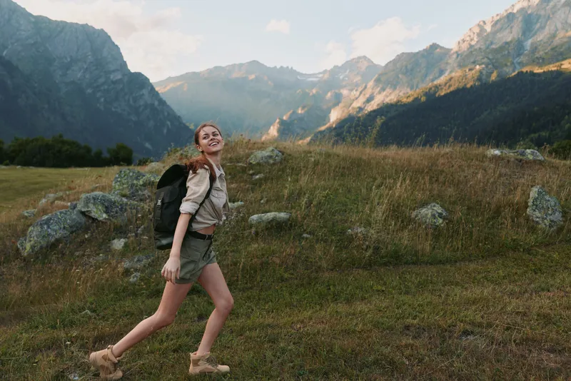 Young woman hiking through a grassy meadow with a mountain range backdrop, wearing a backpack. Emphasizes outdoor activity, nature, adventure, and wellness.