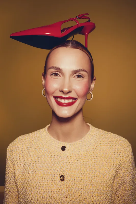 Smiling woman with red high heel shoe balanced on her head wears beige sweater and gold hoop earrings. Concept of fashion, style, creativity, and playful portrait in studio lighting.