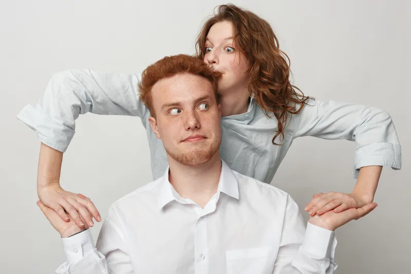 Redhead man and woman in white shirts pose playfully on light background. Woman stands behind man holding his arms with surprised and curious expressions. Concept of friendship and fun.