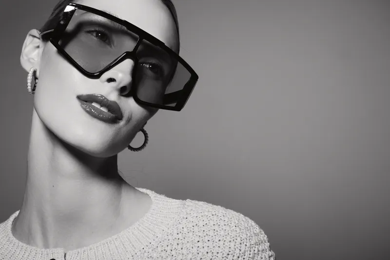 Stylish young white woman with fair skin wearing oversized sunglasses and hoop earrings poses against a plain background. Fashion portrait with modern accessories concept.