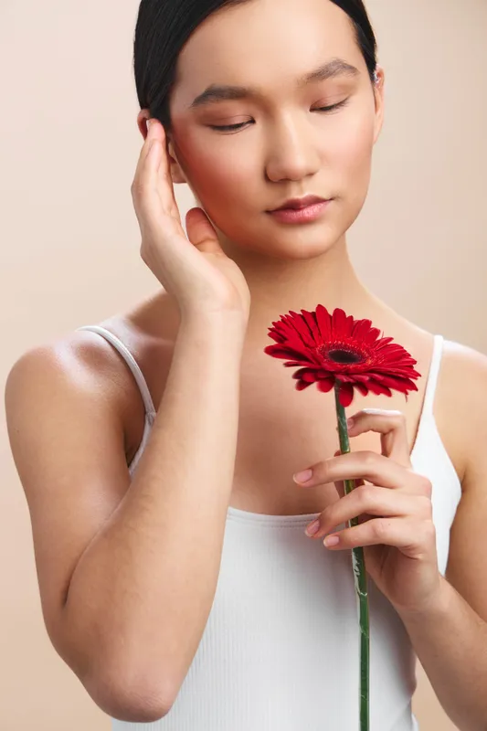 asian woman gently holds a red flower near her face with her eyes closed, conveying calmness and contemplation in a simple studio setting, soft lighting highlighting her serene mood and graceful
