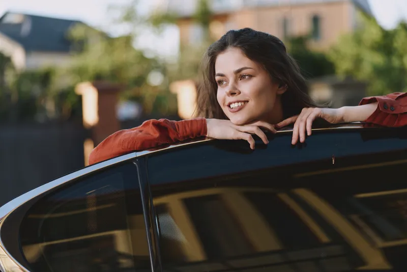 Cheerful woman outdoors, leaning on a car, enjoying a sunlit moment, relaxed posture, gentle breeze, natural colors, urban background, candid lifestyle vibe