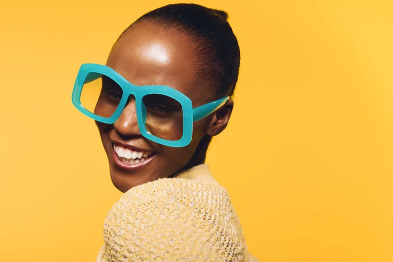 Smiling African woman with dark skin wearing oversized turquoise glasses and beige sweater poses against yellow background. Concept of fashion, joy, and confident beauty portrait.
