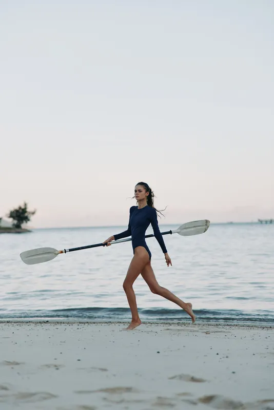 A female paddler in a dark blue wetsuit walks along a calm shoreline, carrying a long paddle, balancing with graceful stride as soft waves touch sandy beach.