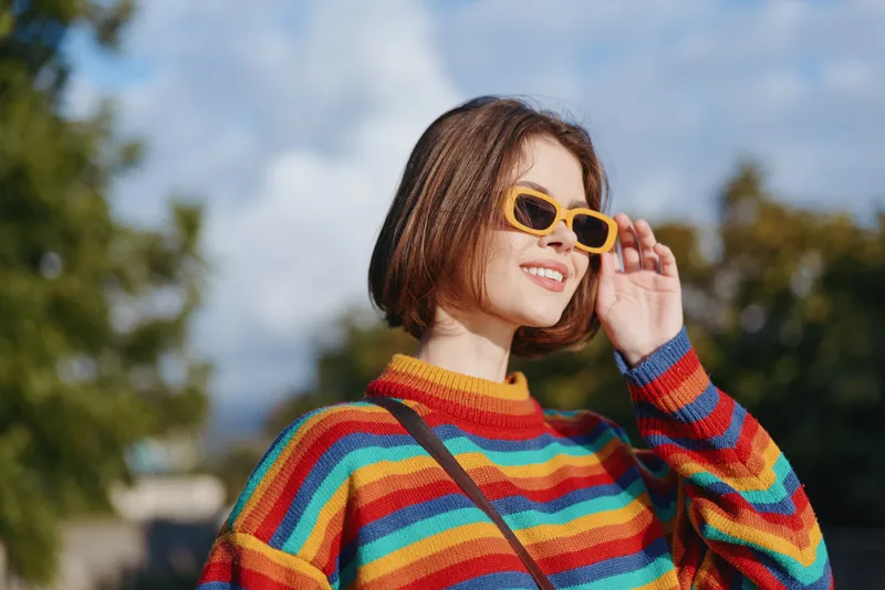 Woman smiling in a rainbow sweater with yellow sunglasses and short hair outdoors, mid twenties model adjusting shades, casual street style, sunny park lifestyle portrait.