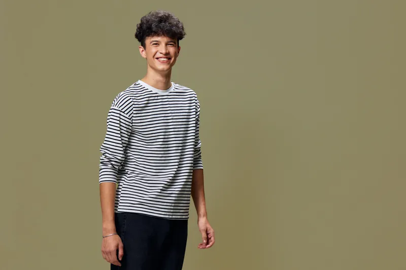 smiling young man wearing striped shirt standing with casual pose against plain olive background, youthful and relaxed expression, indoor studio portrait