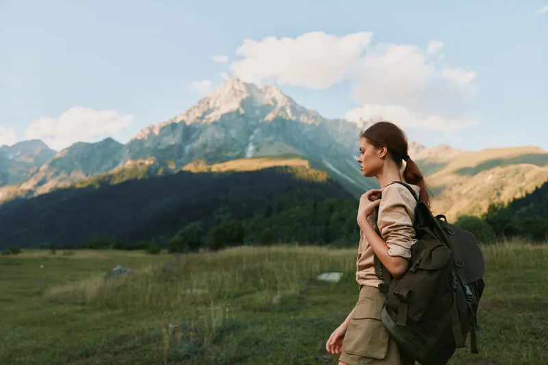 A young woman with a backpack stands in a grassy field looking at a mountain peak under a partly cloudy sky. Concept of outdoor adventure and lifestyle.