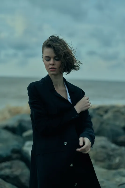 Woman in a dark blazer on a windswept coast, moody fashion scene with sea in the background, elegant solitary posture by the shore, wind swept atmosphere
