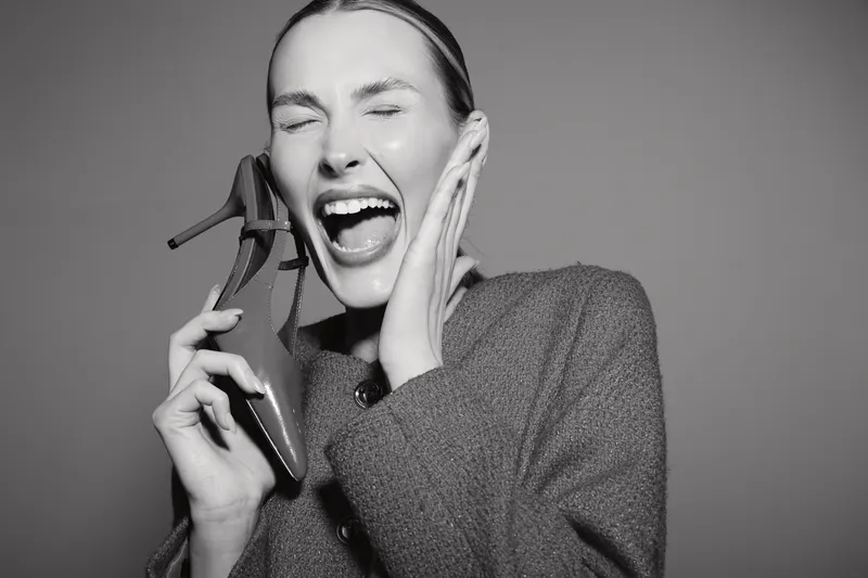 Happy Caucasian woman with light skin laughing while holding a shoe near her ear in a studio with grey background.