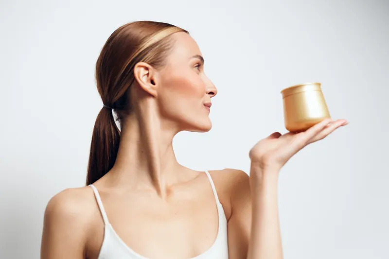 Young woman with light skin in a white tank top holds a small golden jar of cream in profile, testing skincare product against a clean studio backdrop, her expression thoughtful and focused, clear