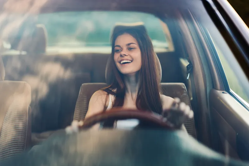 Happy young woman driving a vintage car, enjoying the open road and the sunny weather, with a stylish outfit and a warm smile reflecting carefree moments.