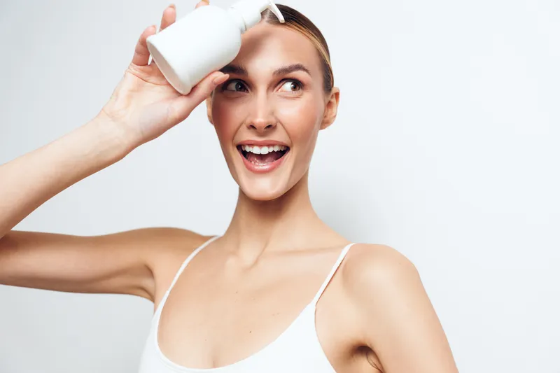 Smiling woman with clear skin holding white bottle near forehead. Studio portrait on white background. Concept of skincare, beauty, health, and self-care with natural light.