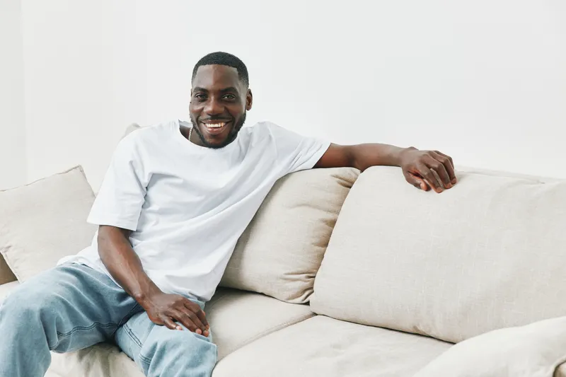Happy African American man in casual outfit sitting on a beige sofa, enjoying a moment of relaxation and positivity in a bright, minimalist living space
