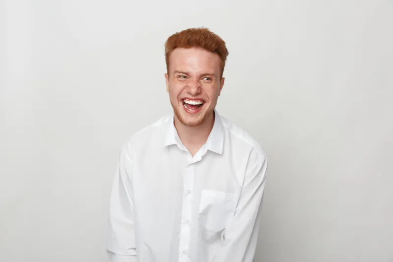 Happy young man with red hair wearing white shirt laughs joyfully against plain background. Concept of positive emotions, youth, and casual lifestyle in studio portrait.