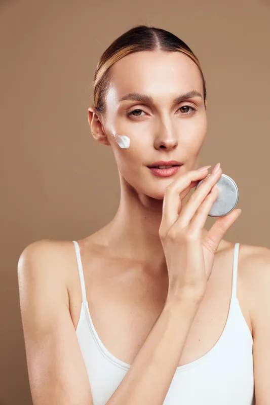 Young woman with natural skin applying cream on face using cotton pad, neutral background, minimal makeup, skincare routine, soft lighting, clean and fresh look