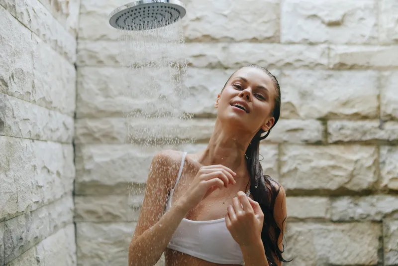 young woman enjoying outdoor shower with water droplets falling on her skin, natural stone background, relaxed mood, summer wellness and freshness