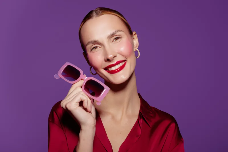 Smiling woman with red lipstick holds pink sunglasses near face on purple background. Fashion portrait with bright makeup and stylish casual outfit. Concept of beauty, style, and confidence.