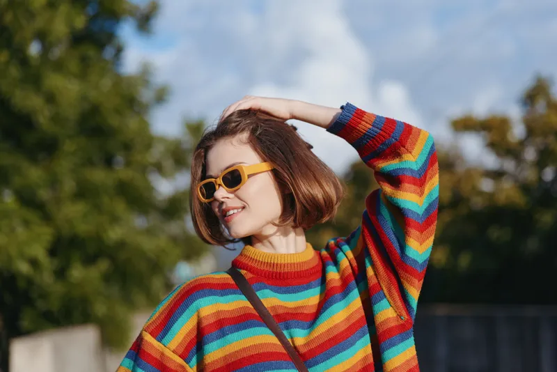 Young woman wearing a colorful striped sweater and yellow sunglasses, smiling with bob haircut in an outdoor portrait, casual fashion lifestyle pose in a sunlit park for travel editorial.