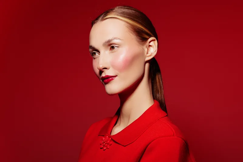 Portrait of a young woman with slicked back hair wearing a red top against a red background. The image highlights makeup, fashion, and beauty concepts with a confident expression.