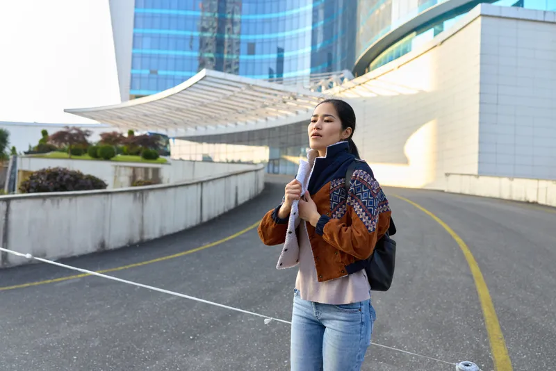 Woman with backpack on a modern city street, scarf wrapped, casual jacket, travel vibe under sunny glass towers, urban scene with road lines and barriers