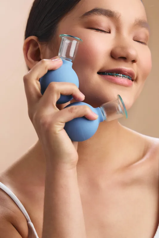 asian woman smiles softly while holding a blue medical device near her cheek, exuding confidence and comfort in a clean studio setting with gentle lighting.