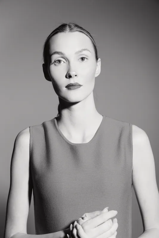 Tall fair skinned white woman with slicked hair wearing sleeveless top looks serious against plain background in black and white portrait.