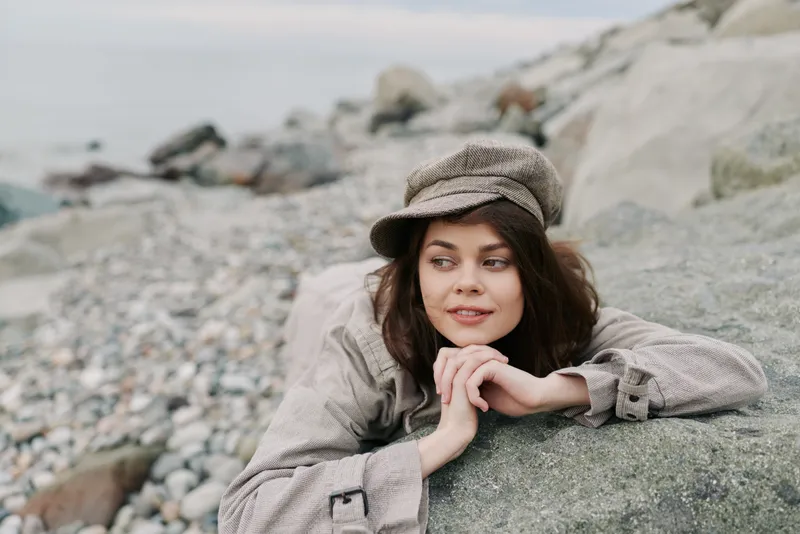 Young woman in casual clothes and hat lies on rocky beach, looking sideways with a relaxed smile. Concept of leisure, nature, and peaceful outdoor moments.