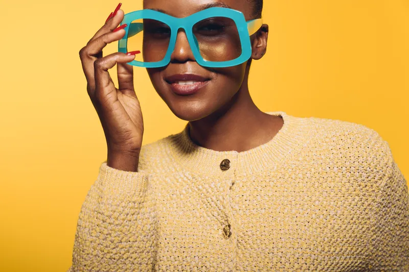 Close-up of a smiling black woman with dark skin wearing large blue glasses and a beige knitted sweater against a yellow background. Concept of fashion, beauty, and confidence.