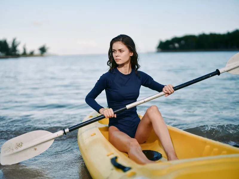 A focused woman paddles a yellow kayak across calm waters, gripping a double?bladed paddle as she glides with determined posture.