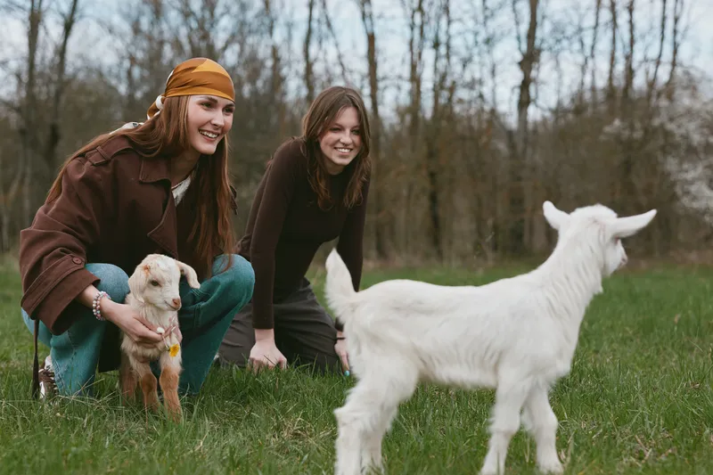 Two friends in casual outfits interact with a goat kid on a grassy pasture, capturing a lifestyle outdoors moment with playful animals and companionship in a natural meadow setting, a candid scene of